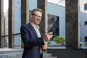A distinguished senior businessman checks his smartphone while enjoying a coffee break outside a contemporary office structure. photo