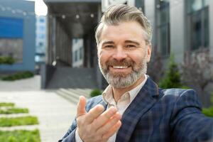 Portrait of a professional senior businessman with a welcoming gesture, standing confidently outside a modern office building. photo