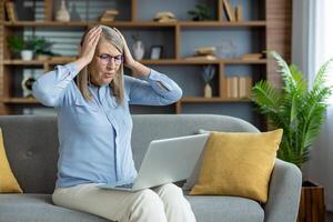 Upset senior woman holding her head, sitting on a couch while using a laptop and experiencing a computer error at home. photo
