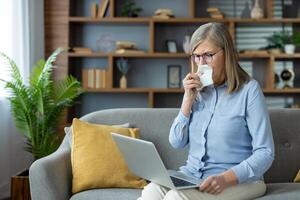 A senior woman crying and using a tissue during an online consultation with a psychologist at home on a laptop. photo