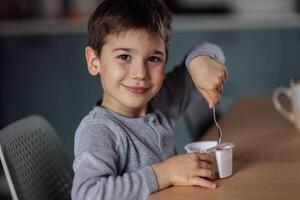 Close up of portrait of cute little boy eats delicious yogurt or cottage cheese from white container with spoon at table in dining room. photo