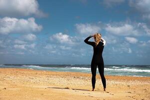 young woman in a wetsuit stands on the beach on the sand against the backdrop of a stormy sea with waves and looks to the side, rear view. Girl in neoprene suit photo