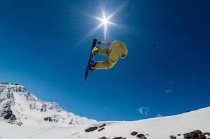 snowboarder jumps from a big air with a board grab, a nose or tail trick in rotation, against the backdrop of snow-capped mountains and the bright sun photo