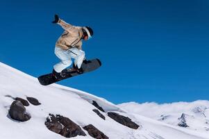 snowboarder jumps into the air against a background of blue sky and mountains. The guy grabs the edge of the board with his front hand while jumping, performing a trick photo