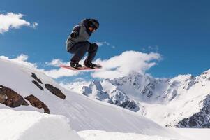 Jumping snowboarder holds one hand on a snowboard against the background of a blue sky and large snow-capped mountains. Performing a stunt in flight photo