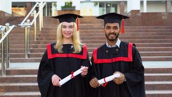 Portrait of happy multiracial graduates. Two friends in graduation caps and gowns standing outside university building, holding diploma scrolls, and smiling video