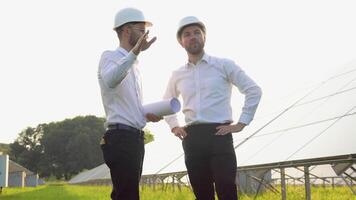 Two men in hard helmets discussing project at solar farm. Male engineers with scrolls of engineering drawing communicating. Concept of green energy video