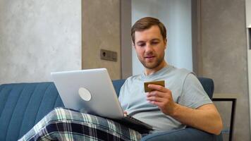Happy Caucasian man buying things online, using laptop and credit card, enjoying shopping in internet. Positive young guy purchasing goods on web, making remote payment video