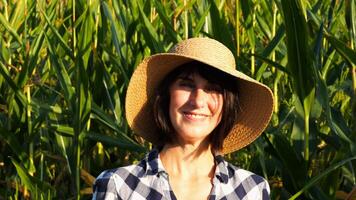 Happy smiling female farmer looks into camera standing near corn field. Portrait of adult beautiful agronomist in straw hat with maize meadow at background. Agricultural business concept photo