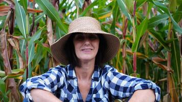 Happy smiling female farmer looks into camera sitting near corn field. Portrait of adult tired agronomist in straw hat with maize meadow at background. Concept of agricultural business. Close up photo