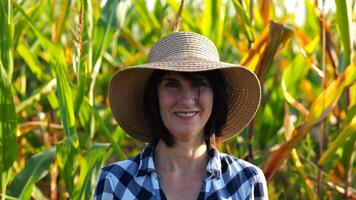 Happy smiling female farmer looks into camera standing near corn field. Portrait of adult beautiful agronomist in straw hat with maize meadow at background. Agricultural business concept. Close up photo
