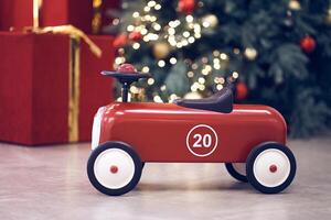 View of red children's retro car on the background of gifts in red boxes with golden bows, Christmas tree decorated garland, string light, red and blue balls. Background, copy space, bokeh photo
