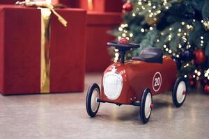 Side view of red children's retro car on the background of gifts in red boxes with golden bows, Christmas tree decorated garland, string light, red and blue balls. Background, copy space, bokeh photo