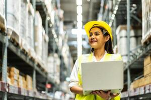Smiling asian woman warehouse worker doing stocktaking of products management in cardboard box on shelves in warehouse using laptop computer, logistic and business export photo