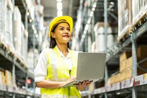 Smiling asian woman warehouse worker doing stocktaking of products management in cardboard box on shelves in warehouse using laptop computer, logistic and business export photo