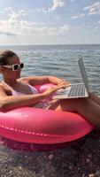 Woman freelancer works on laptop swimming in sea on pink inflatable ring. Happy tourist in sunglasses floating on inflatable donut and working on laptop computer in calm ocean. Remote working anywhere video