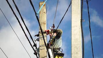 Skilled electrician in helmet fixes wires standing on ladder near high pole against blue sky on summer day backside view. Electrical service and mounting on the pole. Slow motion video