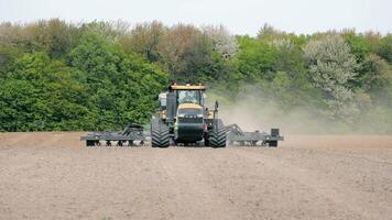 Tractor Preparing Field for Planting, A large tractor equipped with farming machinery plowing a vast field surrounded by dense green forest video