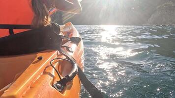 A young girl in an orange kayak paddles through calm waters, the sun reflecting off the surface. The kayak is in the foreground, with the young girl's hand holding the paddle. The sun is shining in video