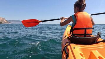 A woman paddles a yellow kayak on a calm blue ocean, wearing an orange life jacket, with a cliff face visible in the distance. video