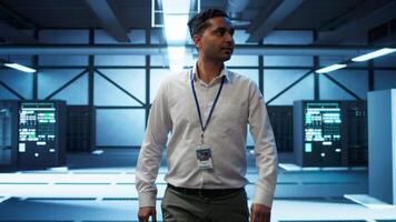 Admin walking amidst server rows, looking to ensure optimal performance and reliability by implement and managing gear. IT staff member in data center overseeing high tech facility, camera B video