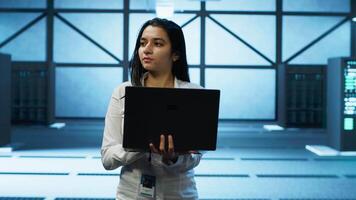 Programmer walking in data center, using notebook to design, implement and manage gear, ensuring optimal performance and reliability. IT professional in server hub coding on laptop, camera A video