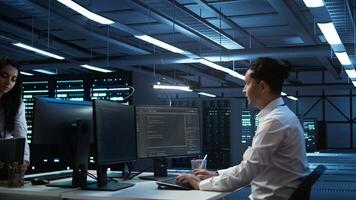 Team of programmers in high tech data center troubleshooting issues using tablet. Teamworking colleagues in server farm using device, establishing performance benchmarks, camera B video