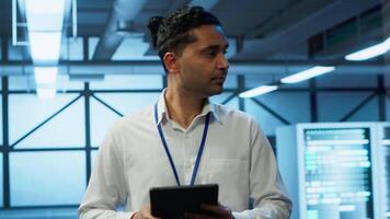 Programmer walking in high tech data center used for managing infrastructure energy consumption, working on tablet. Indian man using device in workspace used for automation in server farm, camera A video