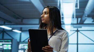 Software developer walking in data center, using tablet to design, implement and manage gear, ensuring optimal performance and reliability. IT worker in server farm coding on device, camera A video