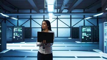 Programmer walking in data center, using notebook to design, implement and manage gear, ensuring optimal performance and reliability. IT professional in server hub coding on laptop, camera B video