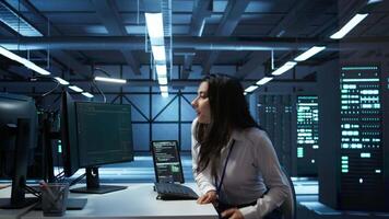 Engineer in data center sitting down at desk, installing software on computer, troubleshooting issues. Woman arriving at work in server hub, establishing performance benchmarks, camera B video