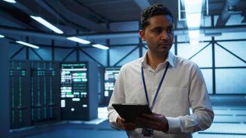 Programmer walking in high tech data center used for managing infrastructure energy consumption, working on tablet. Indian man using device in workspace used for automation in server farm, camera B video