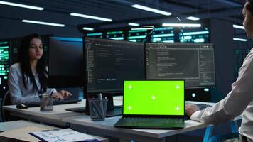 Zoom in on green screen laptop used by data center worker troubleshooting issues. Engineer uses chroma key notebook in server hub, establishing performance benchmarks, camera B video