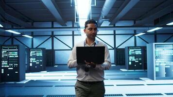 Technician using laptop in data center, managing servers, operating systems. Professional in server hub using notebook to ensure networks are secure and functioning properly, camera B video
