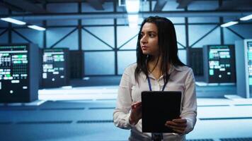 Software developer walking in data center, using tablet to design, implement and manage gear, ensuring optimal performance and reliability. IT worker in server farm coding on device, camera B video