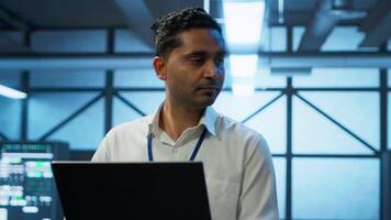 System administrator in data center using notebook to design, implement and manage gear, ensuring optimal performance and reliability. Indian man in server room coding on laptop, camera A video