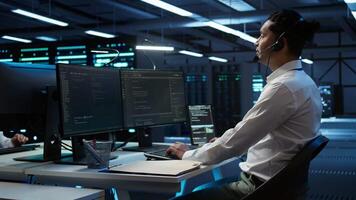 Data center computer scientist wearing mic headset, doing installation of networking equipment. Server hub IT professional talking in microphone with system administrators, camera B video