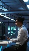 Vertical System administrator in data center installing software on computer, troubleshooting issues using tablet. IT staff member in server hub establishing performance benchmarks, camera B video