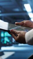 Vertical Data center admin responsible for installation of networking systems solving tasks in workspace. Close up of tablet used by IT worker doing maintenance on server farm gear, camera B video