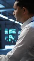 Vertical Portrait of smiling technician in data center implementing and managing infrastructure to ensure optimal performance and reliability. Happy IT specialist in server room coding on computer, camera B video