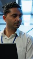 Vertical System administrator in data center using notebook to design, implement and manage gear, ensuring optimal performance and reliability. Indian man in server room coding on laptop, camera A video