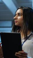 Vertical Software developer walking in data center, using tablet to design, implement and manage gear, ensuring optimal performance and reliability. IT worker in server farm coding on device, camera A video