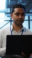 Vertical System administrator in data center using notebook to design, implement and manage gear, ensuring optimal performance and reliability. Indian man in server room coding on laptop, camera B video