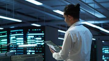 System administrator overseeing data center, making improvements and updates to ensure system can handle networking tasks. IT expert in server room tracking network traffic with tablet, camera A video