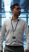 Vertical Admin walking amidst server rows, looking to ensure optimal performance and reliability by implement and managing gear. IT staff member in data center overseeing high tech facility, camera B video