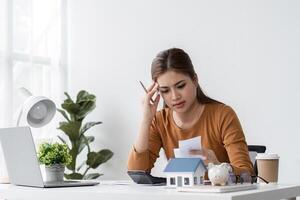 Young Woman Stressed While Selling a House, Working at Desk with Laptop, Documents, and Model House photo