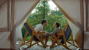 Lovers are kissing while relaxing on the veranda in front of their tent at the campsite, sitting on chairs. Medium shot. Slow motion. video