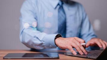A man in a blue shirt and tie is typing on a laptop. He is using a pen to write on a tablet. Concept of professionalism and focus, as the man is working on a task that requires concentration photo