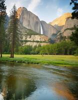 A tranquil river flows through a valley with a majestic waterfall cascading down a towering cliff photo