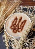 Coffee cup with foam patriotic pattern coat of arms of Ukraine, stencil decor latte mug standing on table with spikelets. latte cappuccino. vertical, top view photo